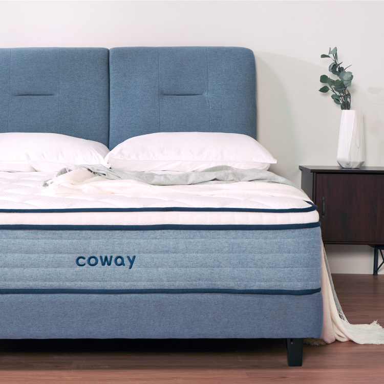 Front View Of The Coway Prime Lite Series Mattress With Complete Bedsheets And A Blue Bedframe Along A Side Table With A Plant Vase  - Coway Prime Lite Series